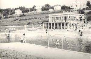 Czarno-białe zdjęcie otwartego basenu kąpielowego. W głebi budynek