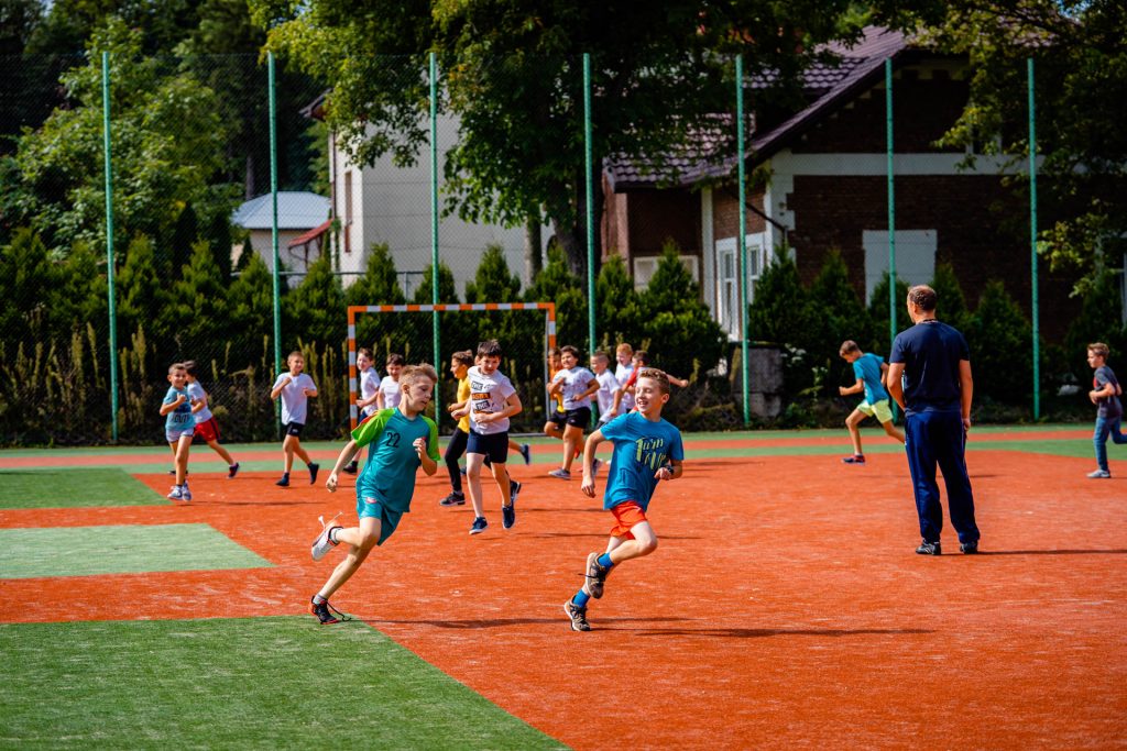Chłopcy w strojach sportowych biegaja po boisku. Nawierzchnia ma kolor ceglasty i zielony