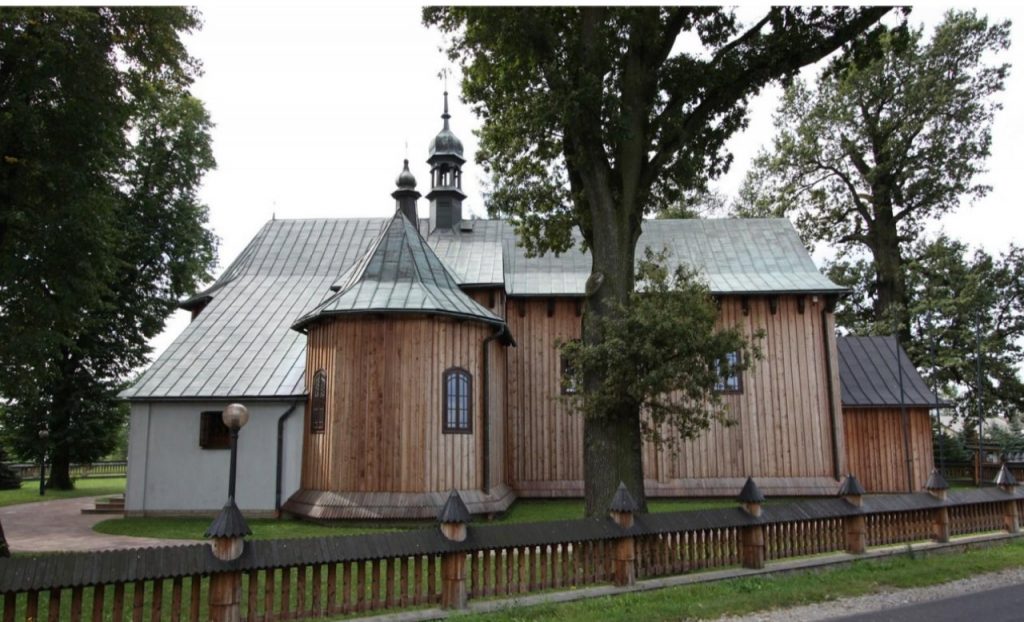 Zabytkowy kościół z jasnego drewna, kryty blaszanym dachem. Przed kościołem duże drzewa liściaste.